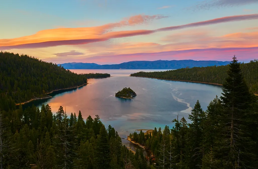 Sunset over a calm lake with a small island, framed by dark pine trees and colorful clouds