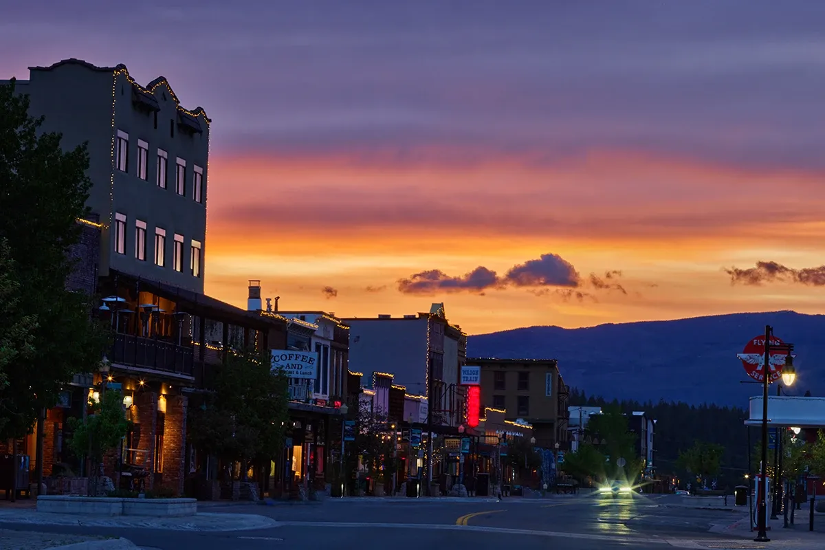 Downtown street at sunset with glowing storefronts, mountains in the distance, and a colorful orange sky