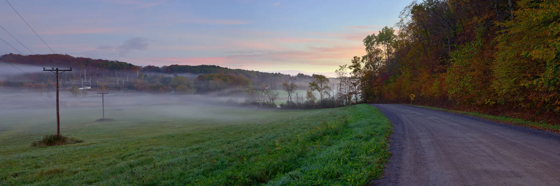Foggy country road curving past a green field at sunrise, with trees and a pink-blue sky