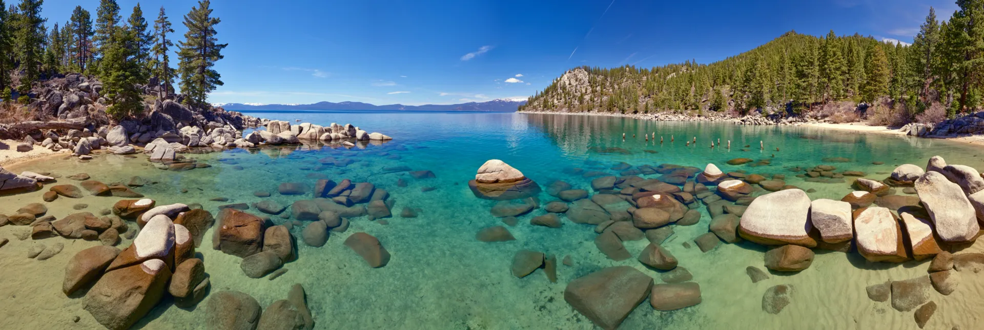 Clear turquoise lake with rocks in the foreground, pine trees along the shore, and mountains under a blue sky.