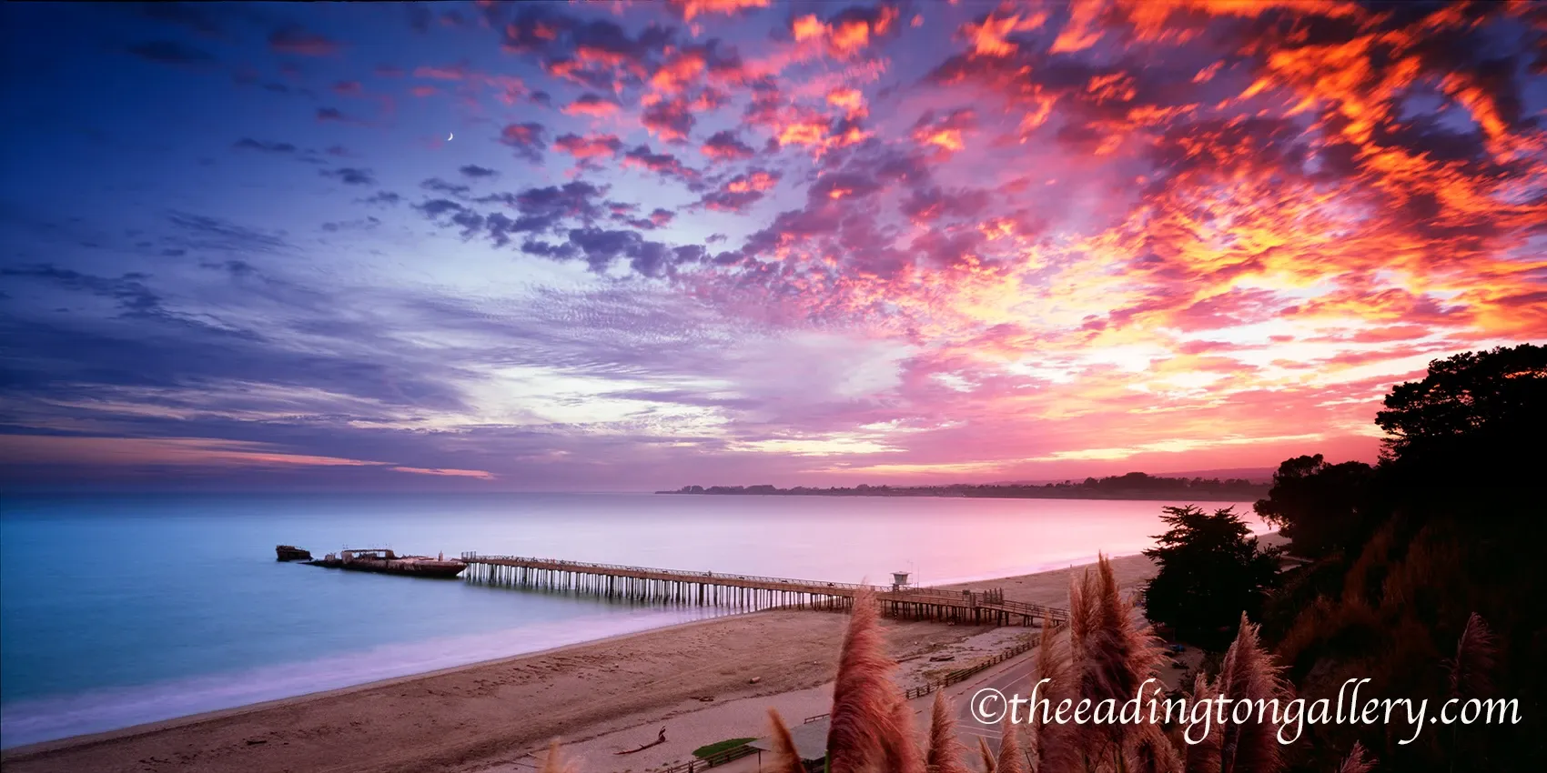 Sunset over a calm beach with a wooden pier and vivid pink-orange clouds