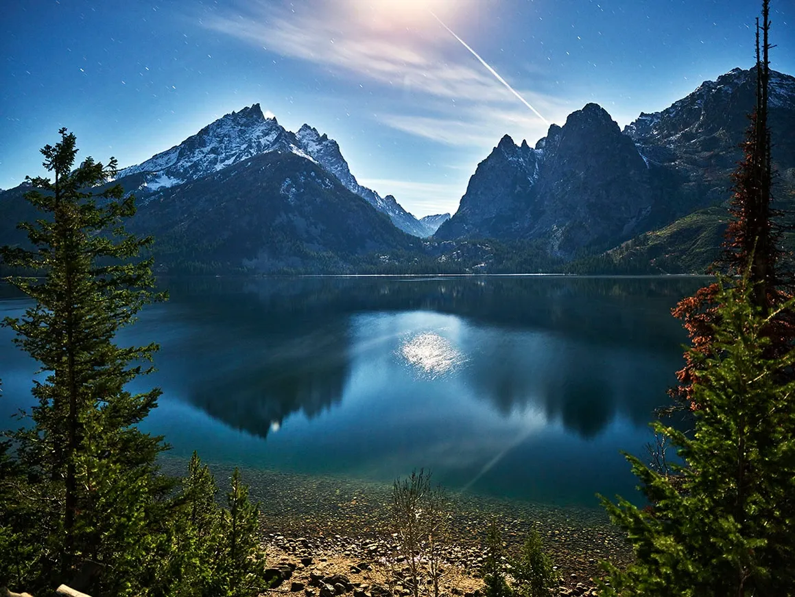 Snowy mountain peaks reflected in a calm lake at sunrise, framed by evergreen trees.