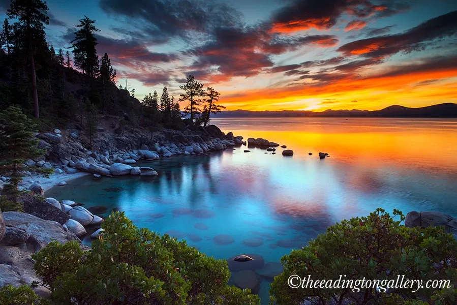 Sunset over a calm blue lake with rocky shore, pine trees, and colorful clouds reflected in the water