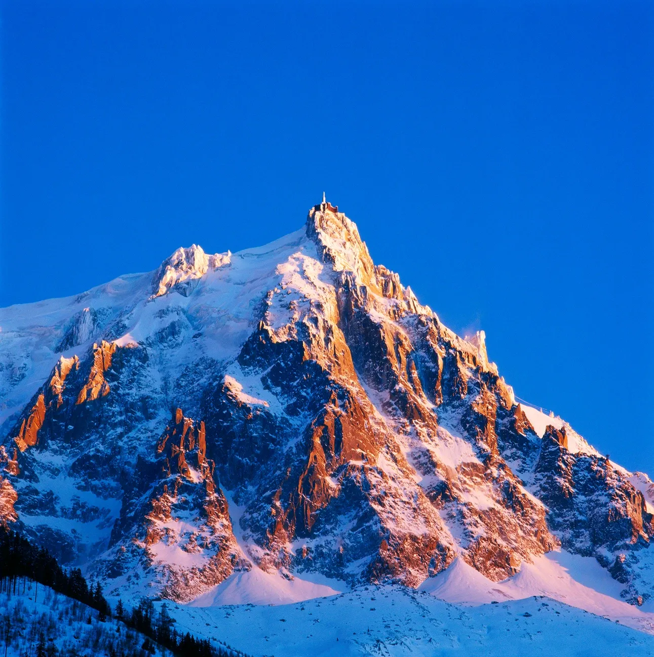Snowy mountain peak lit by warm sunset against a clear blue sky