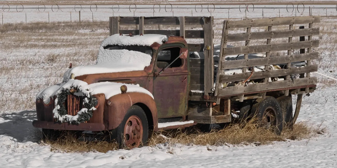 Snow-covered old rusty pickup truck in a field, with a wooden livestock bed.