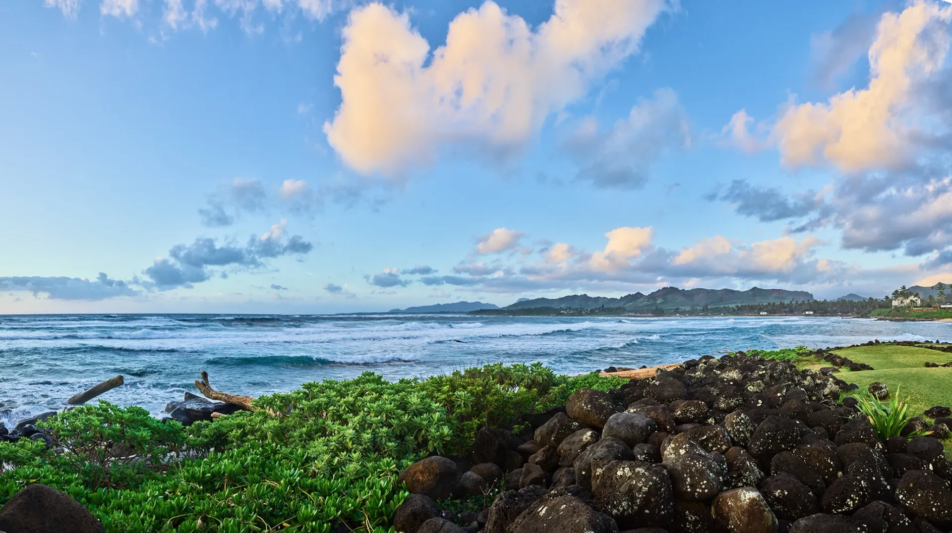 Rocky shoreline with green vegetation, ocean waves, and a partly cloudy blue sky at sunset