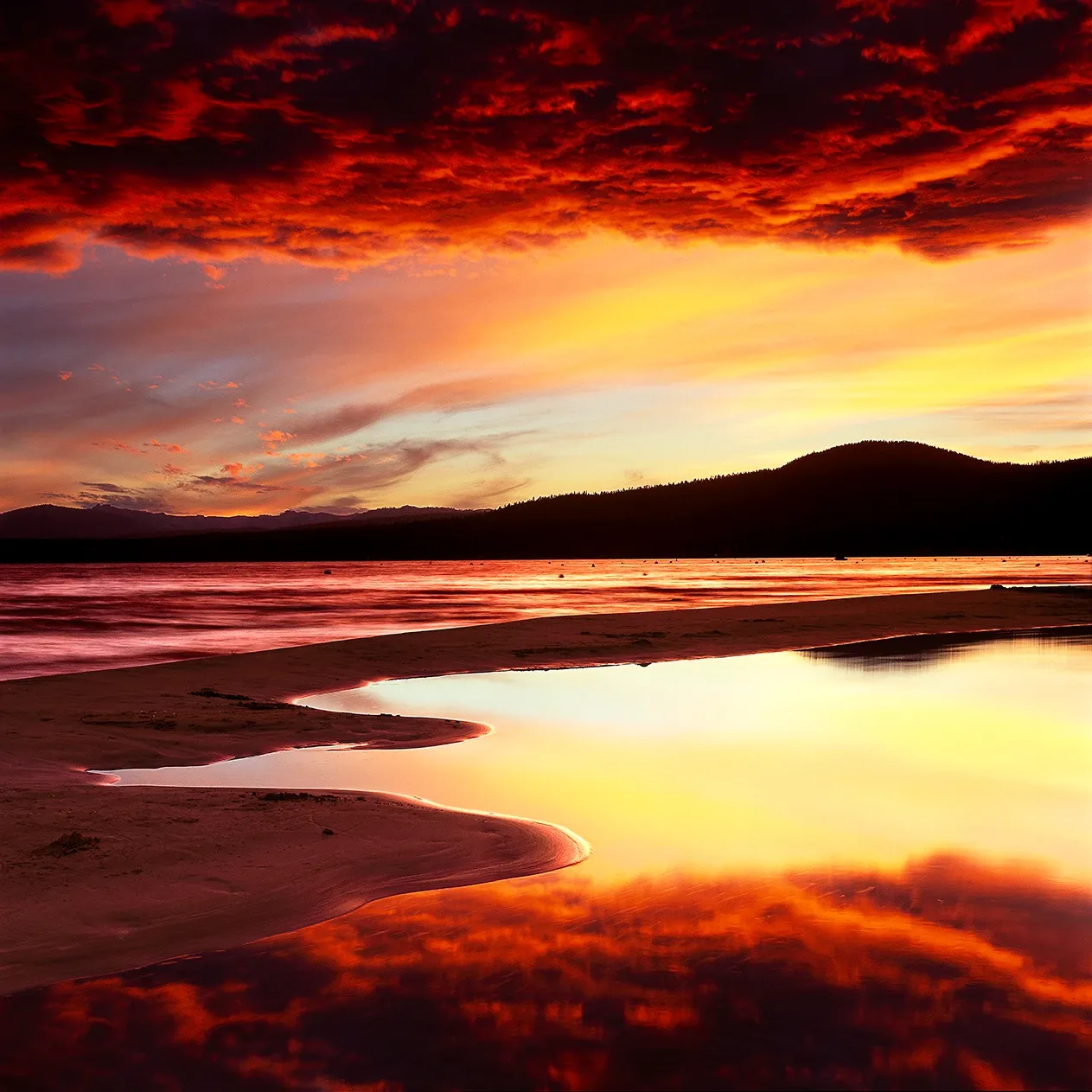 Fiery red-orange sunset over a calm beach with reflective wet sand and dark shoreline silhouette