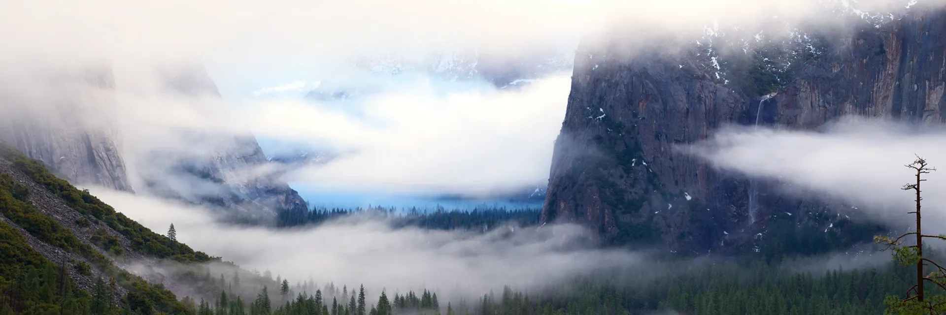 Cloudy mountain valley with low fog, forested slopes, and a blue river or lake in the distance