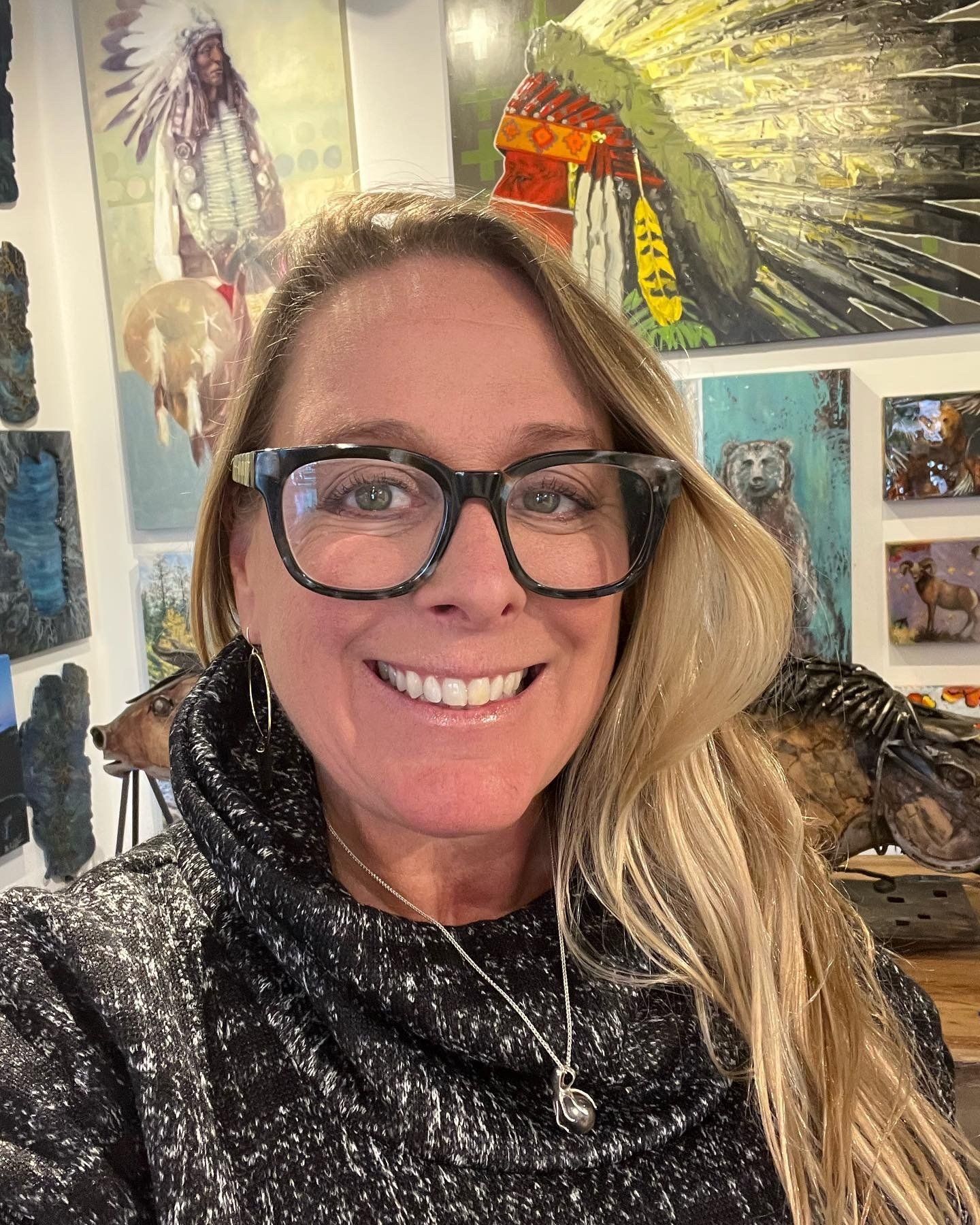 Smiling person with glasses and long blonde hair in front of colorful bird artwork and paintings