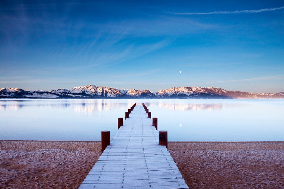 Wooden pier extending into a calm lake with snow-capped mountains and a blue sky at sunset