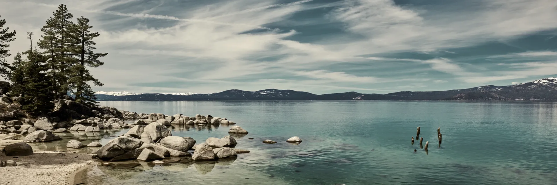 Rocky lakeshore with pine trees and people swimming in calm blue water under a cloudy sky