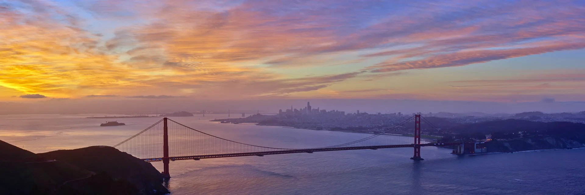 Golden Gate Bridge at sunset over San Francisco Bay, with purple-orange sky and city lights.
