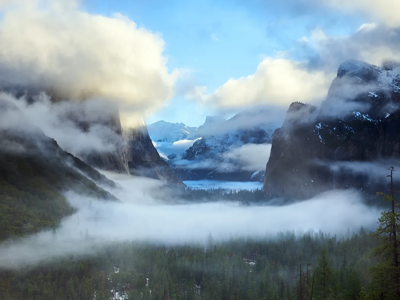 Foggy mountain valley with clouds, dark cliffs, and distant snowy peaks under a blue sky
