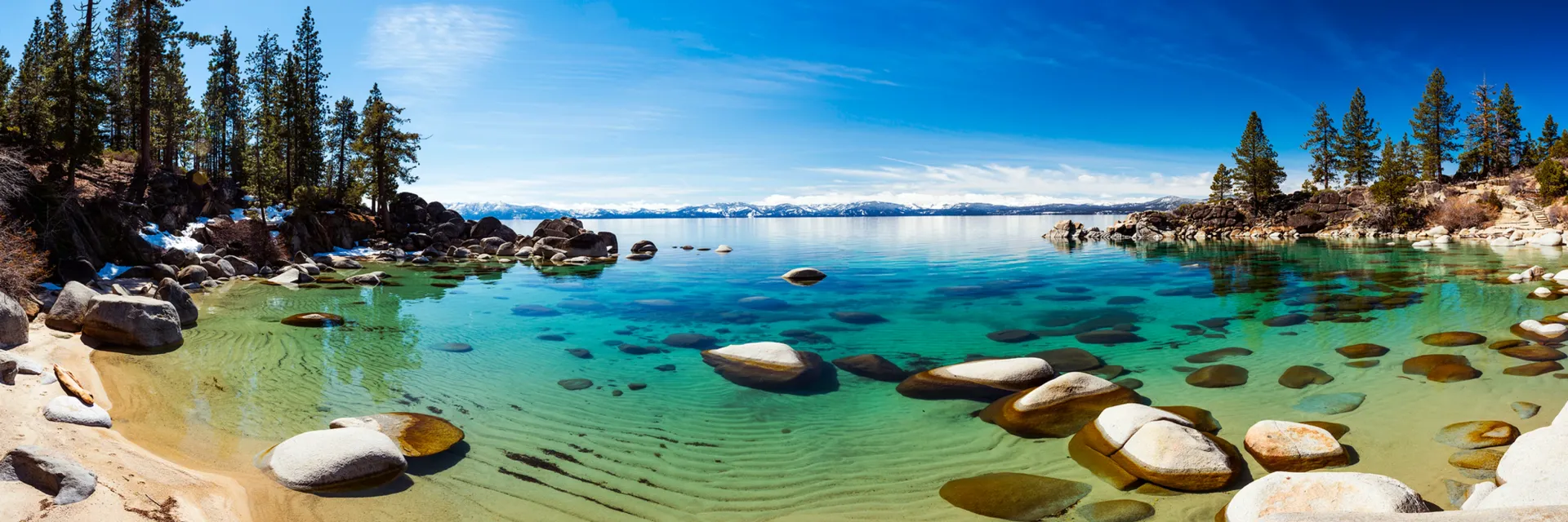 Turquoise mountain lake with rocks, sandy shore, pine trees, and a clear blue sky