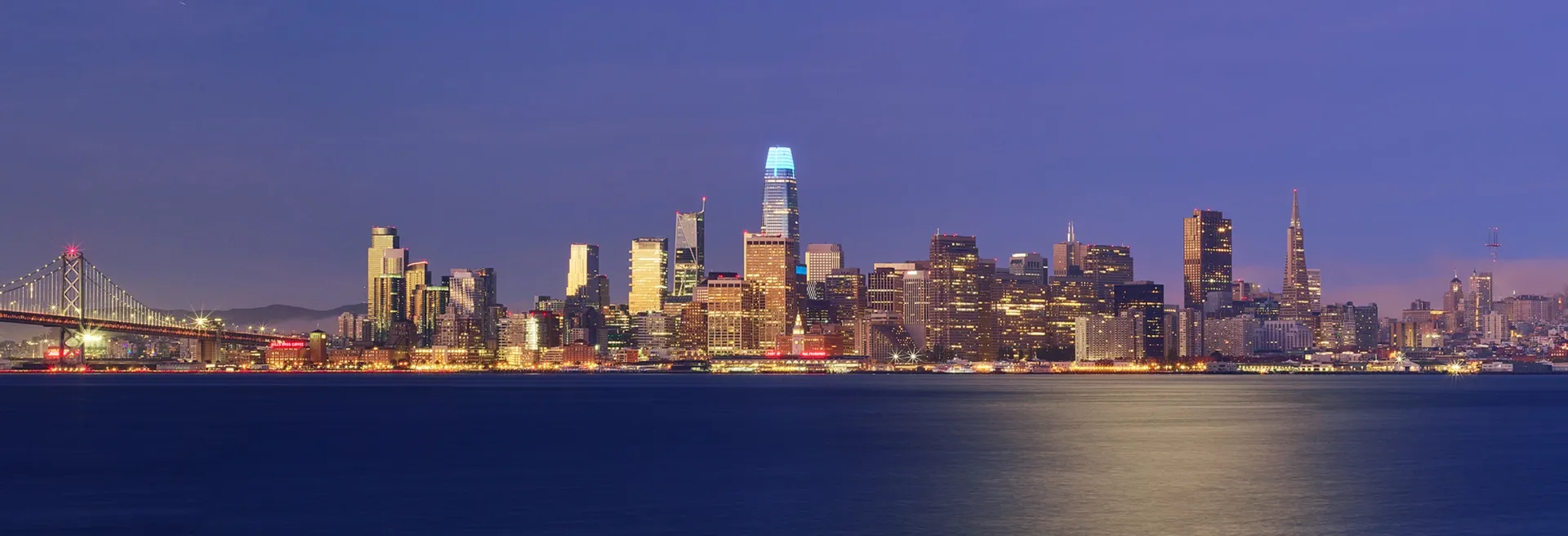 City skyline at dusk across calm water, with lit skyscrapers and a blue-purple sky.