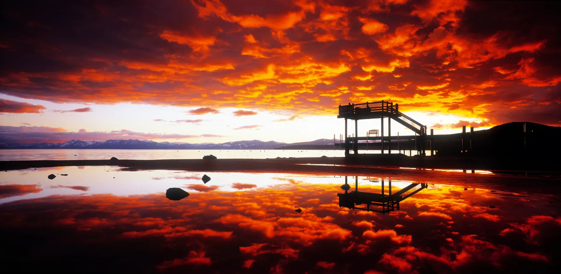 Fiery sunset over calm water with silhouetted dock structures reflected in the shore