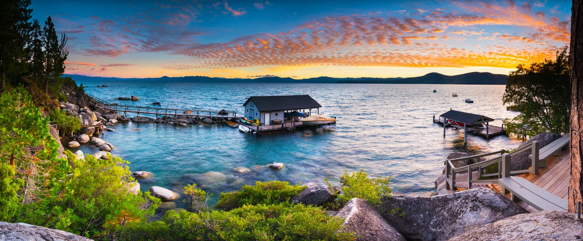Sunset over a rocky lakeside with a small boathouse, dock, and calm water reflecting orange clouds