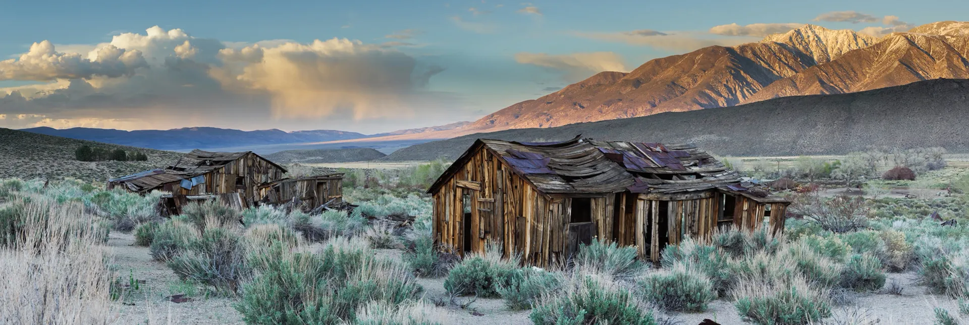 Abandoned cabin in frosty grassland with snow-dusted mountains and cloudy sunrise sky
