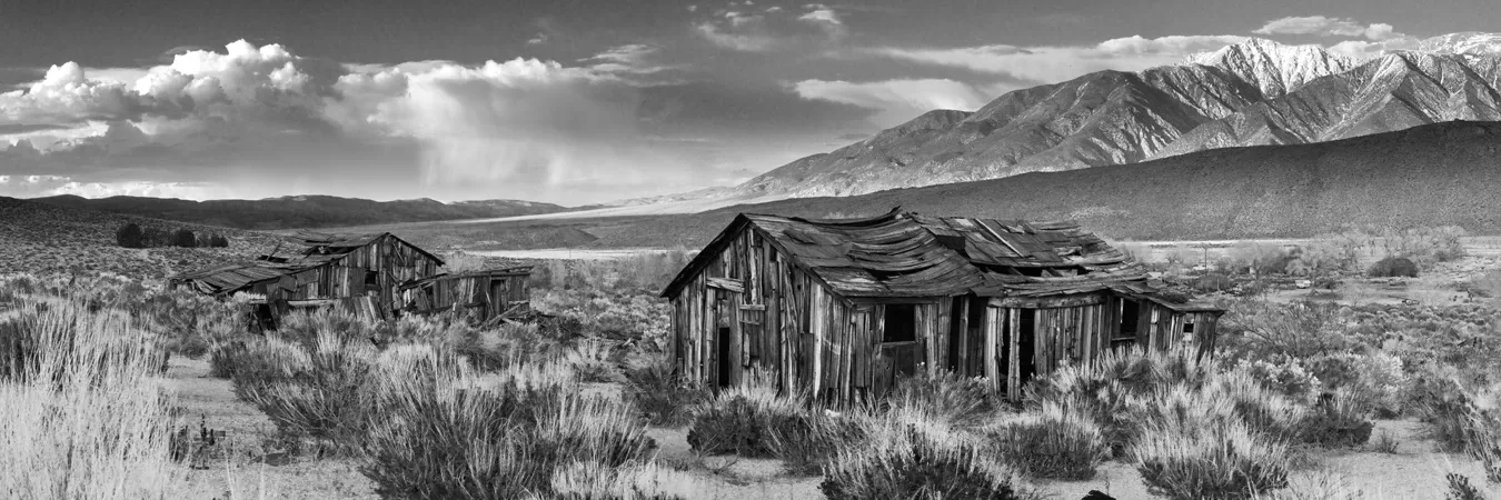 Black-and-white landscape of an abandoned wooden cabin in a grassy plain with mountains and cloudy sky beyond