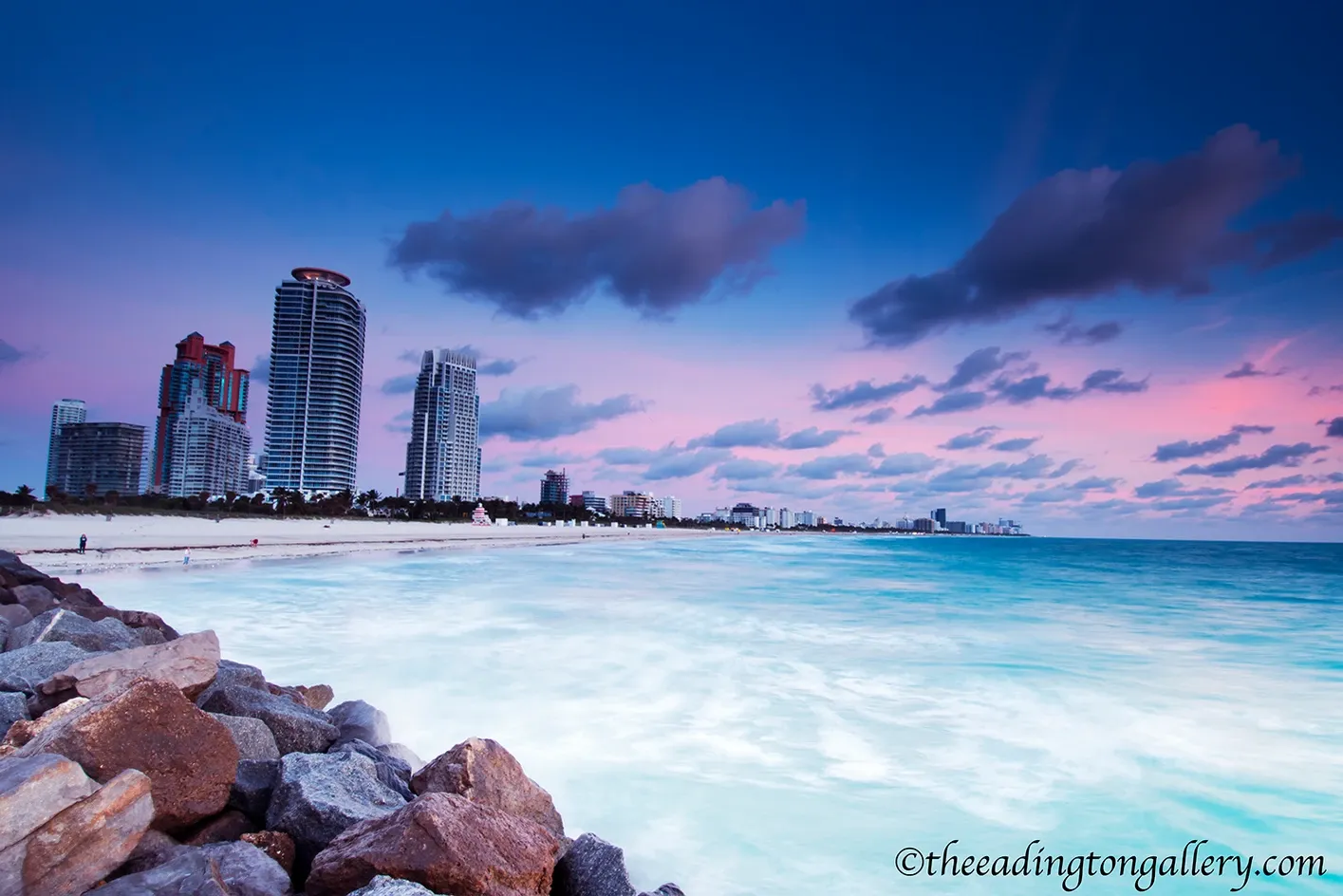 Rocky shoreline with turquoise water, pink dusk sky, and distant city skyscrapers.