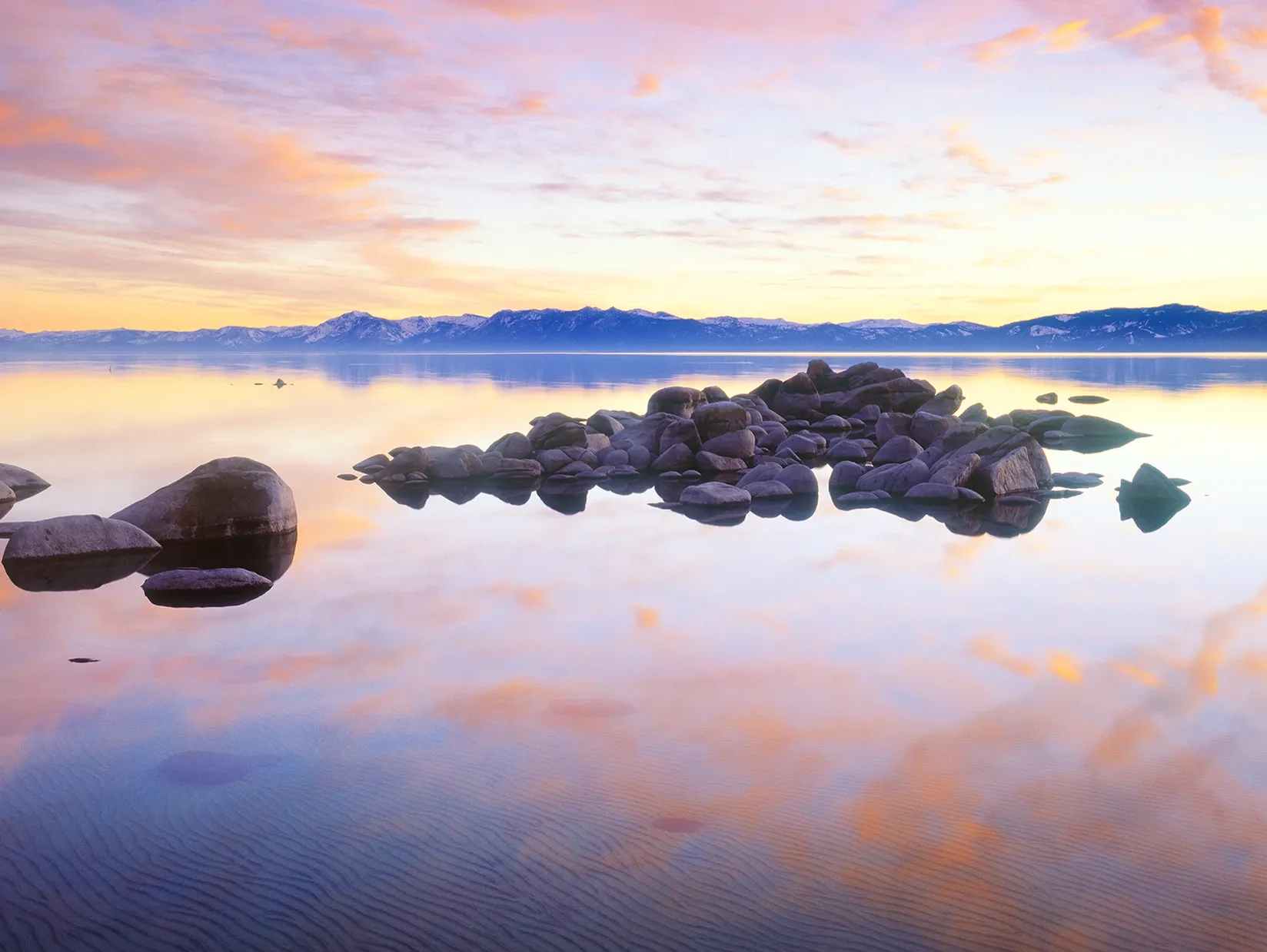 Calm lake at sunrise with mirrored pink sky, distant mountains, and rocks in the foreground
