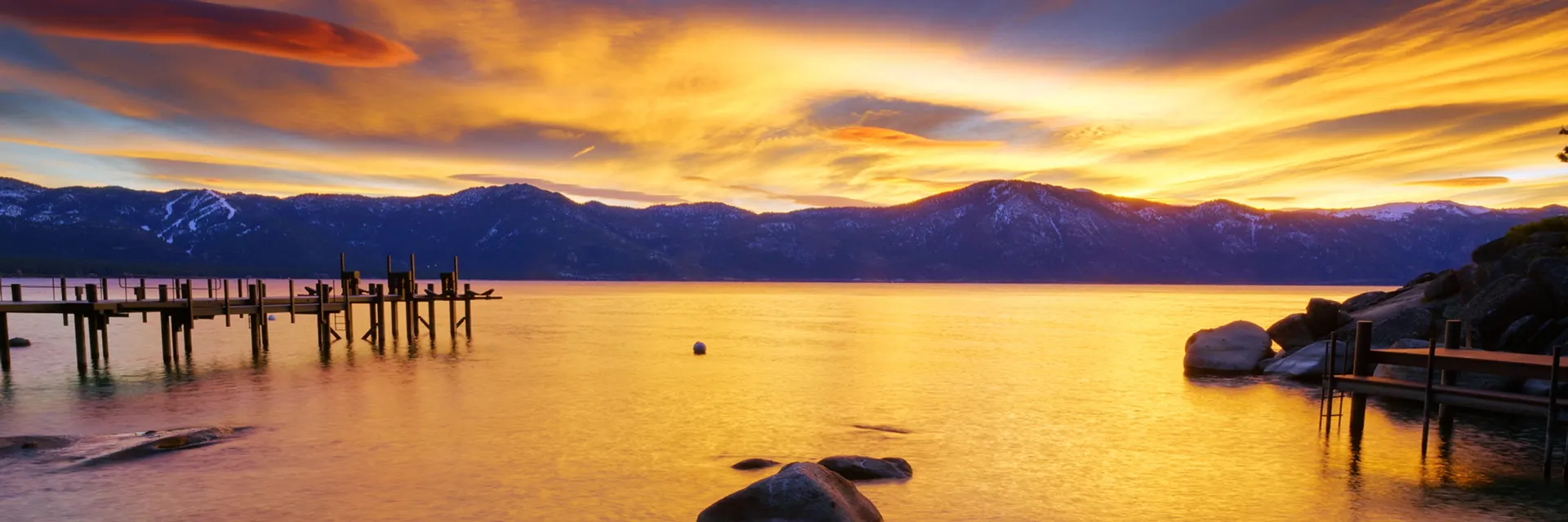 Sunset over a calm lake with orange reflections, mountains in the distance, and a wooden pier on the left