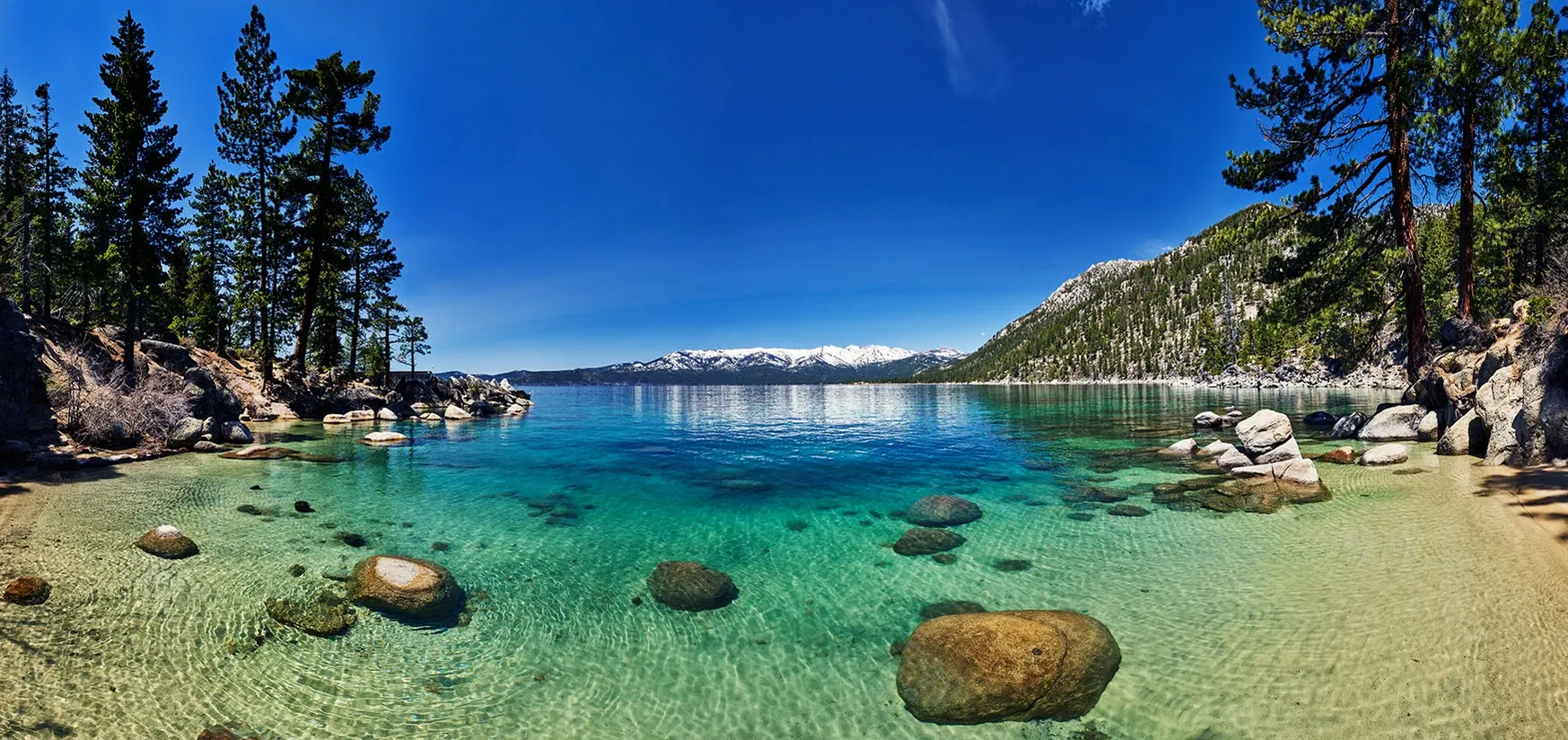 Clear turquoise lake with rocky shore, pine trees, and snow-capped mountains under a blue sky