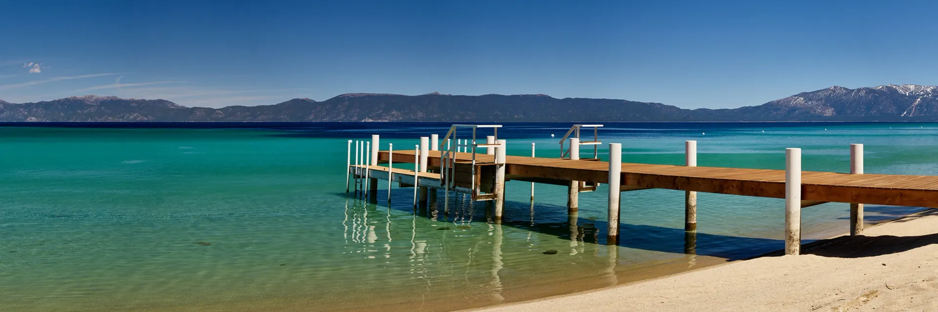 Wooden dock extending into clear turquoise water beside a sandy beach under a blue sky.