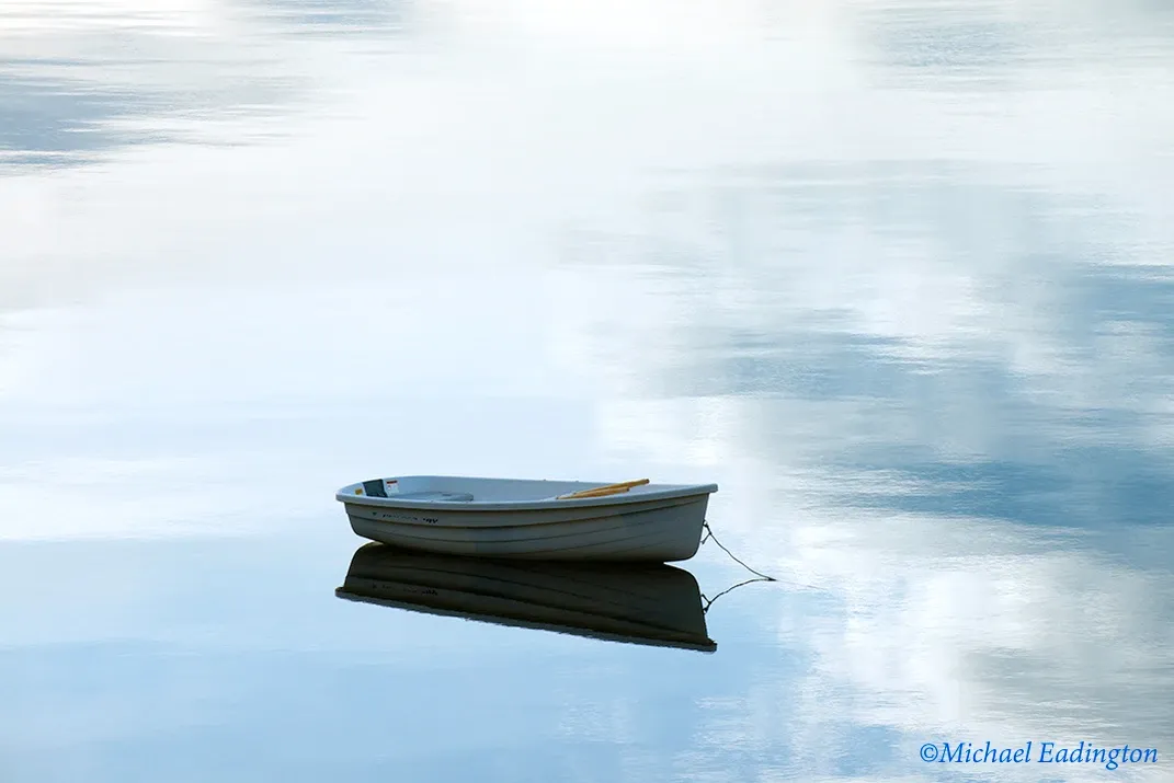 Small white rowboat floating on calm blue water with its reflection visible