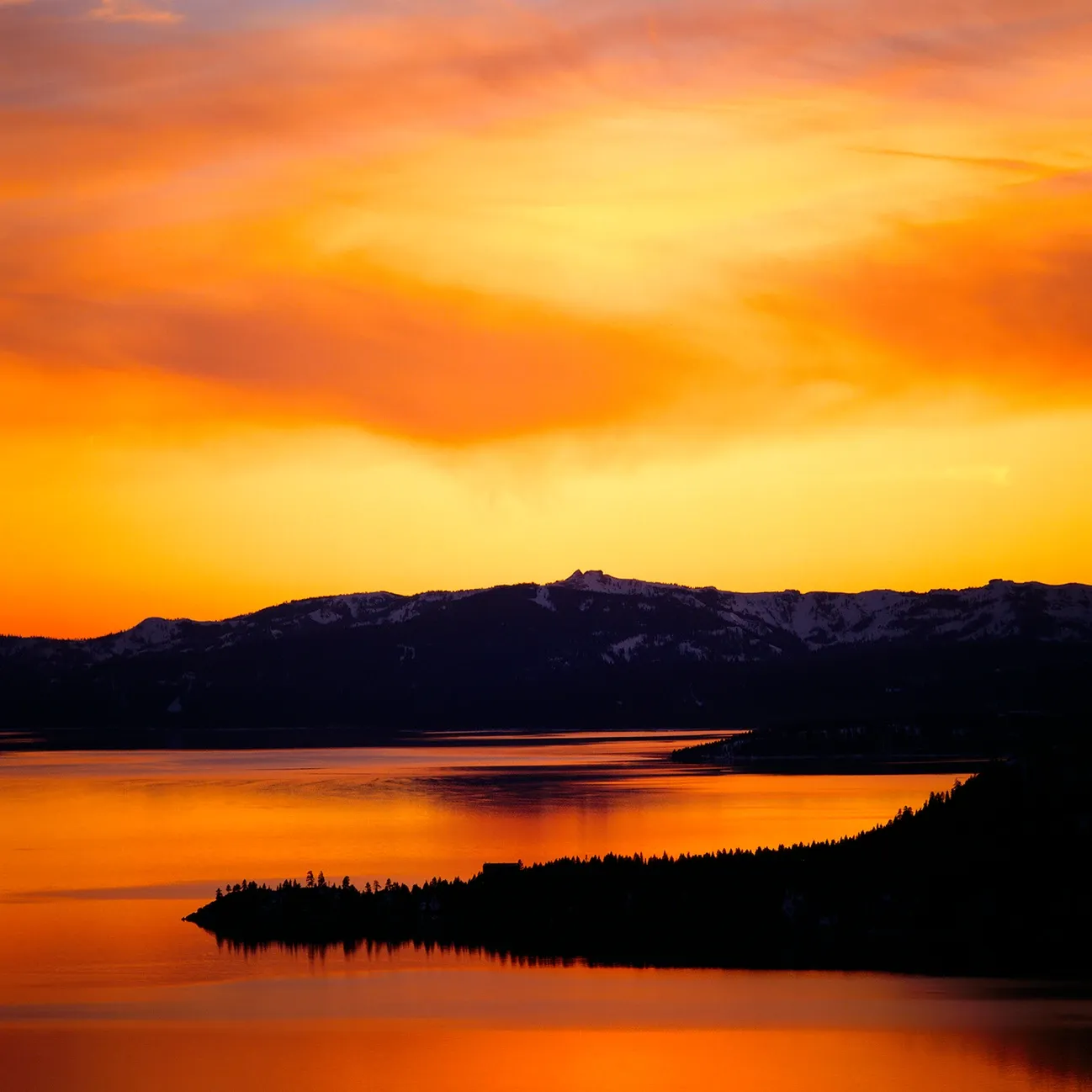 Orange sunset over a calm lake with dark hills and a small tree-covered peninsula