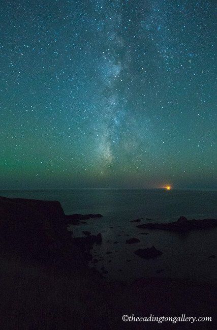 Starry night sky over a dark coastline, with the Milky Way rising above calm water and a distant orange light.