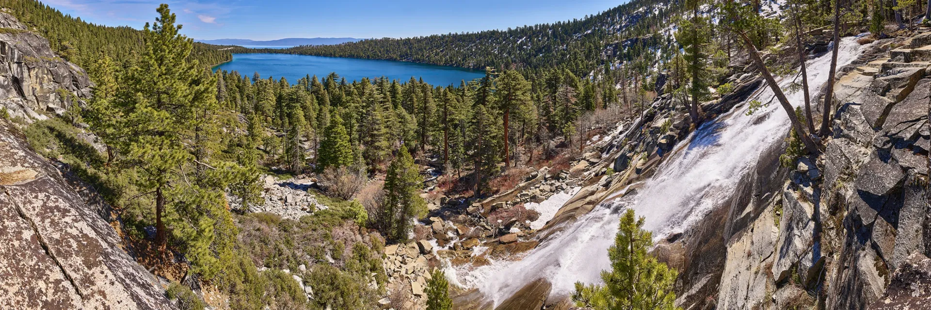 Forest-lined mountain lake with a waterfall cascading down a rocky cliff under a blue sky
