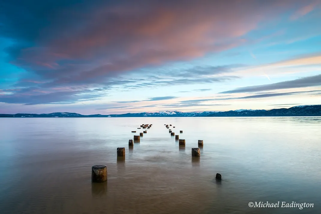 Calm shoreline at dusk with wooden posts leading into still water under a pink-blue sky