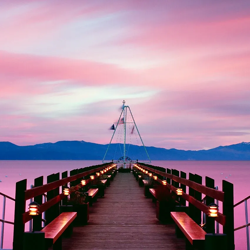 Wooden pier with benches leading to a sailboat on calm water under a pink sunset sky