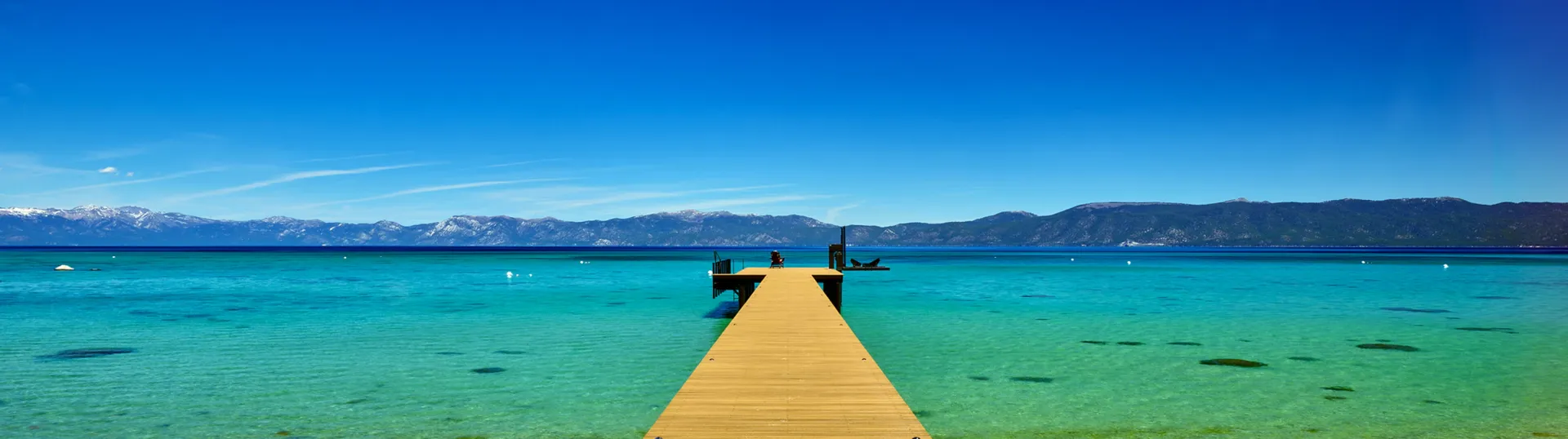 Wooden pier extending into turquoise ocean under a bright blue sky, with distant mountains on the horizon