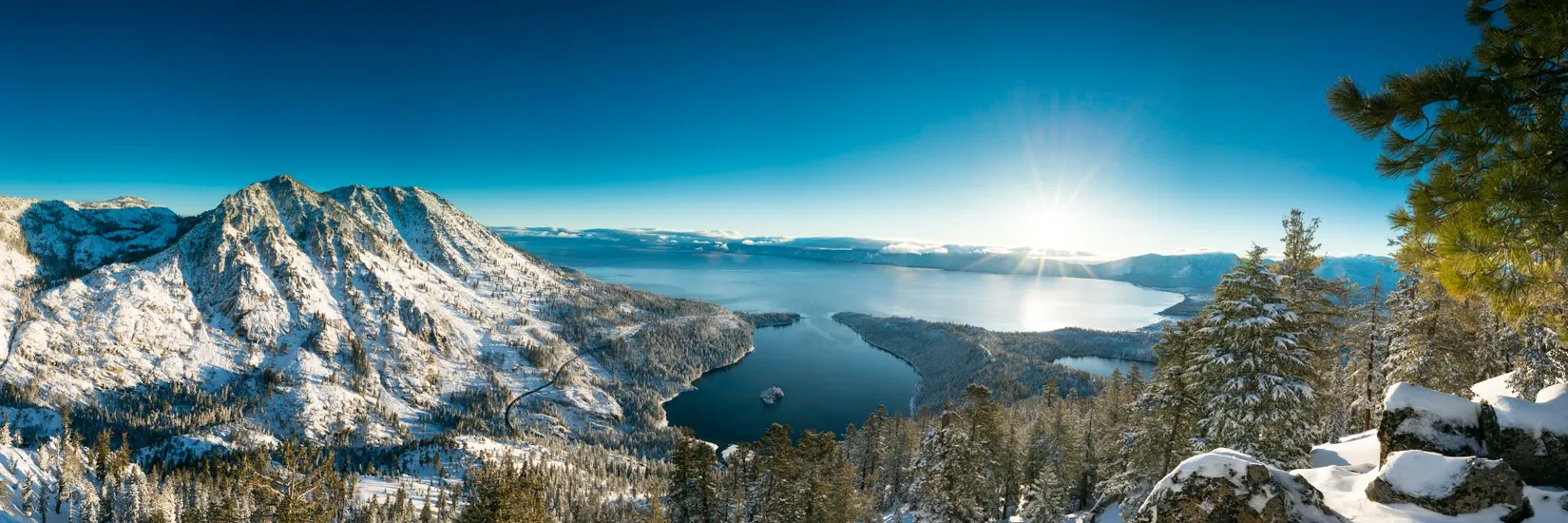 Snowy mountain overlook with a lake, pine trees, and bright blue sky at sunrise