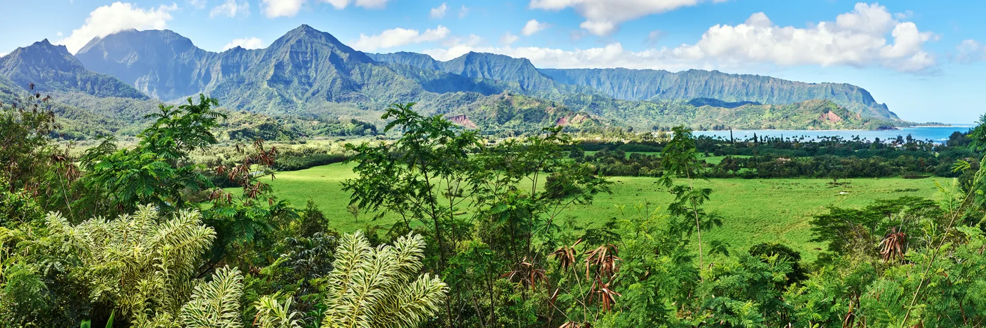Lush green valley with fields and trees beneath rocky mountains under a bright blue sky.