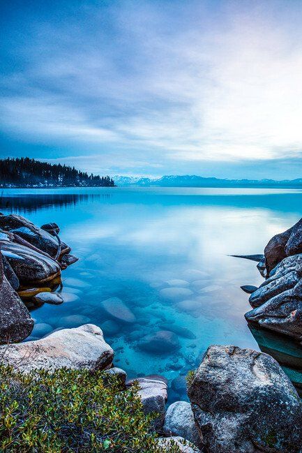 Rocky lakeshore with clear turquoise water, distant pine trees, and a cloudy blue sky