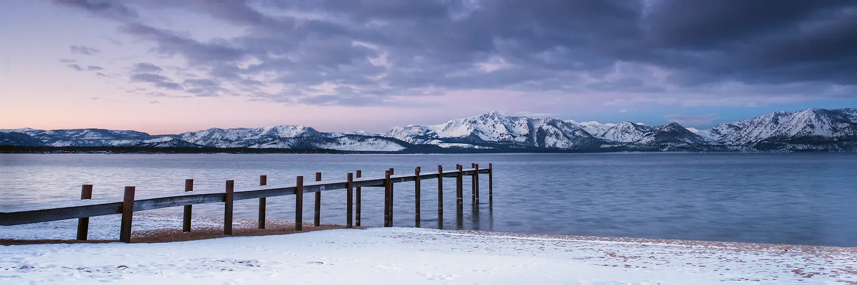 Snowy mountains reflected in a calm lake beside a wooden pier at dusk