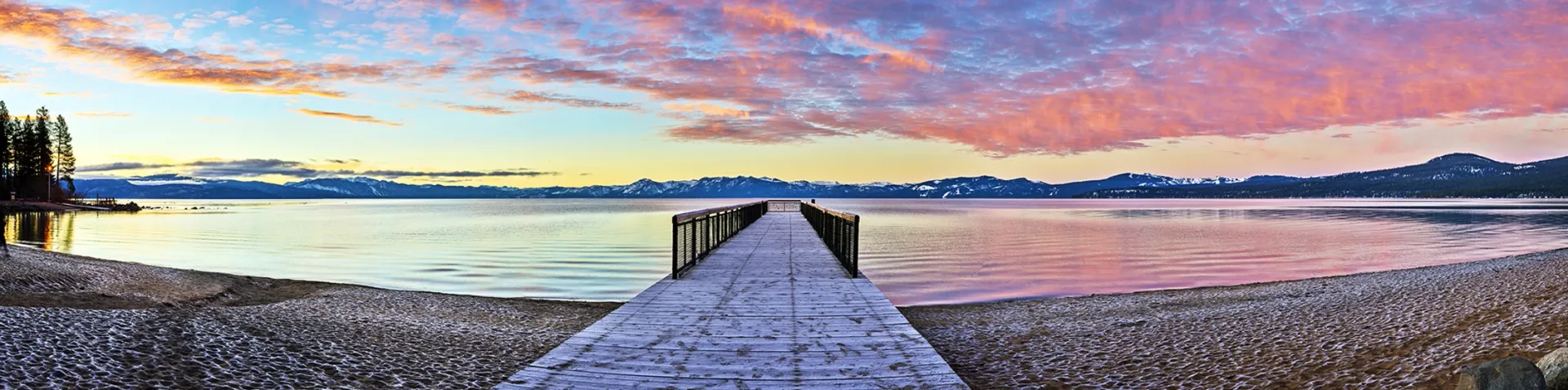 Snow-covered dock leading to a calm lake at sunset, with pink and blue clouds overhead