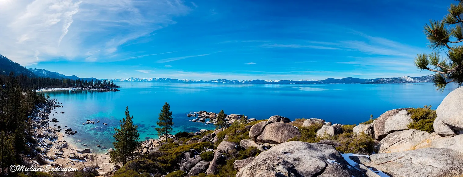Rocky lakeshore with clear blue water, distant mountains, and bright sky under scattered clouds