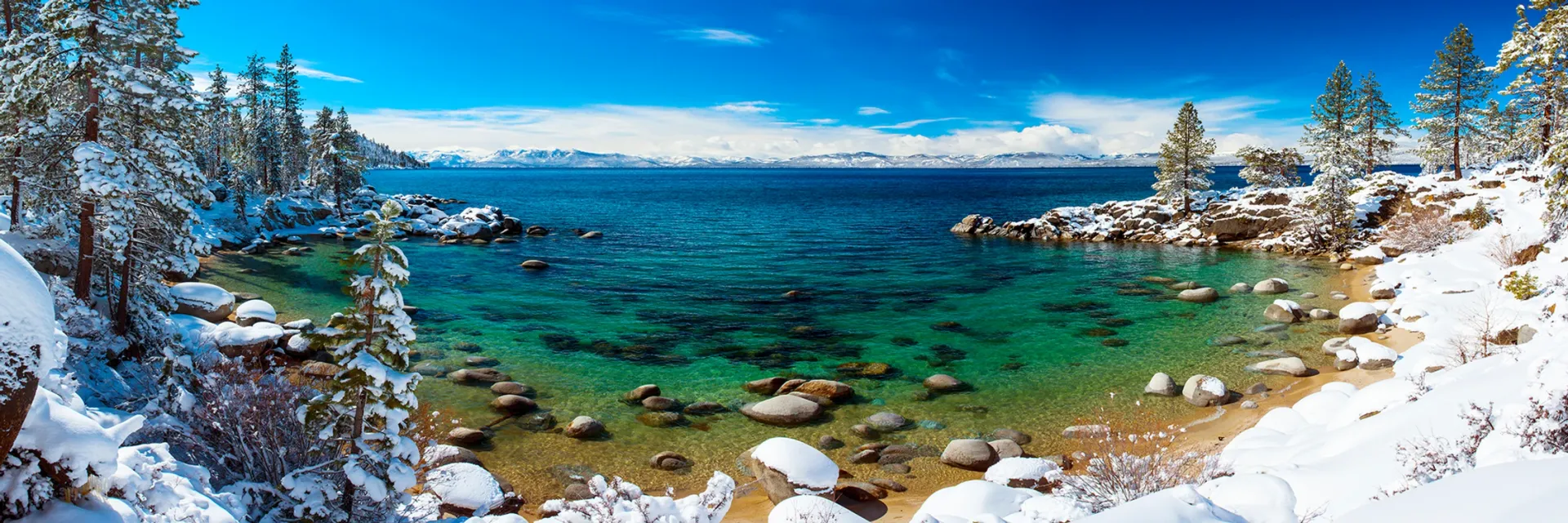 Snowy lakeshore with turquoise water, pine trees, and blue sky under bright sunlight