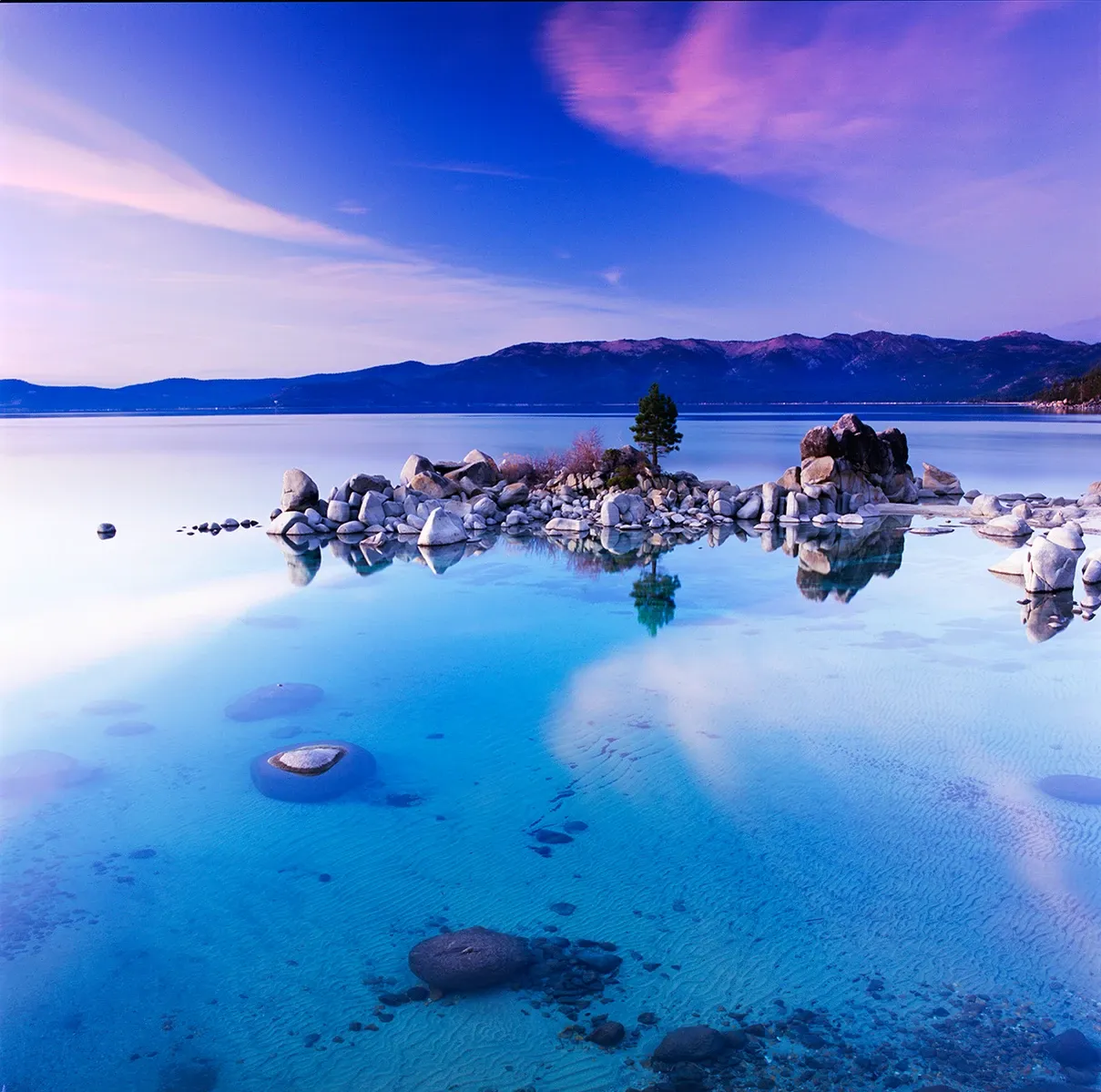 Turquoise lagoon with rocky islet and distant mountains under a pink-blue sky