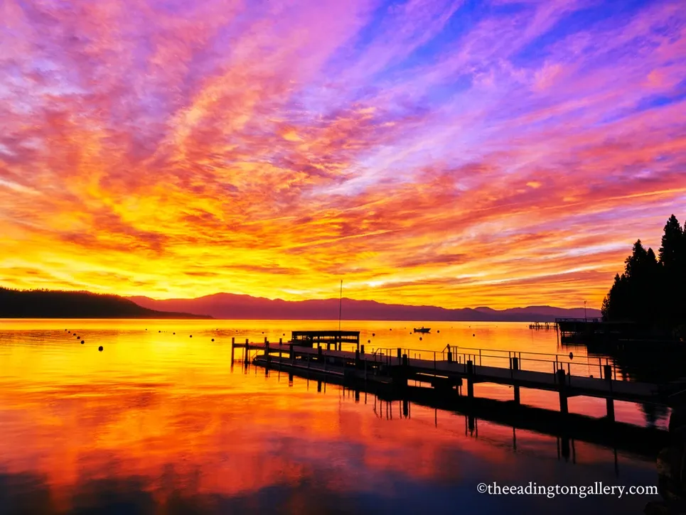 Vibrant orange and purple sunset over a calm lake with a dock and silhouetted mountains