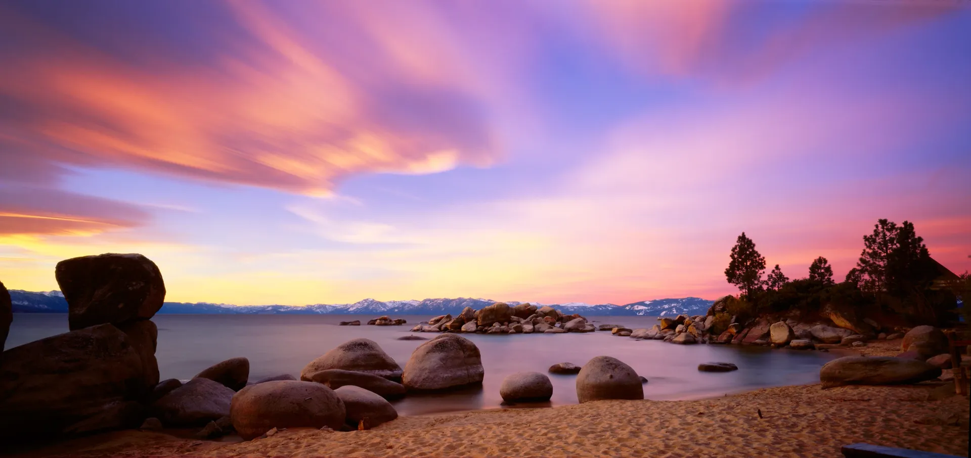 Sunset over a rocky beach with calm water, silhouetted trees, and pink-purple clouds