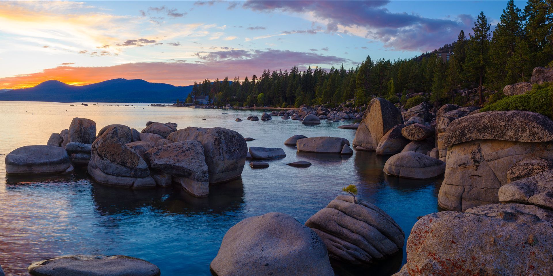 Rocky coastline at sunset with calm blue water and distant mountains under a colorful sky