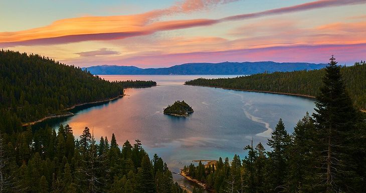 Sunset over a calm blue lake with a small island, framed by dark pine forests and orange-pink clouds