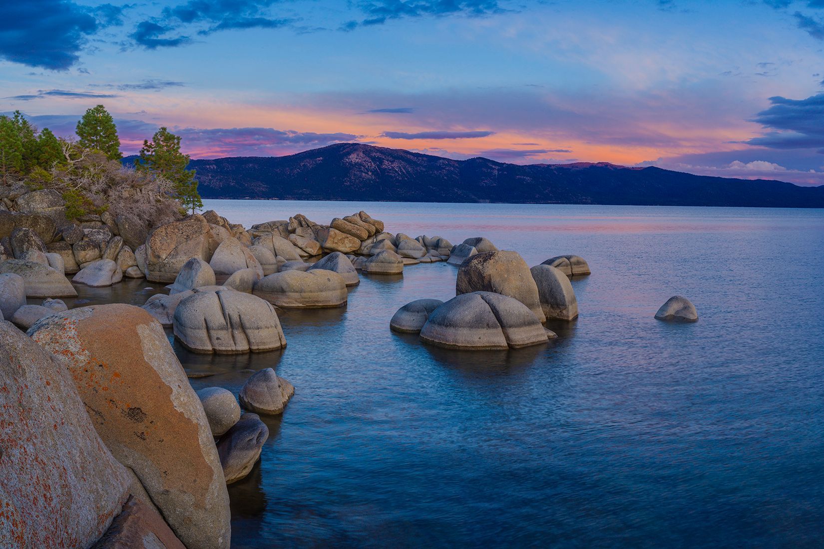 Rocky lakeshore at dusk with calm blue water and pink-purple sunset sky