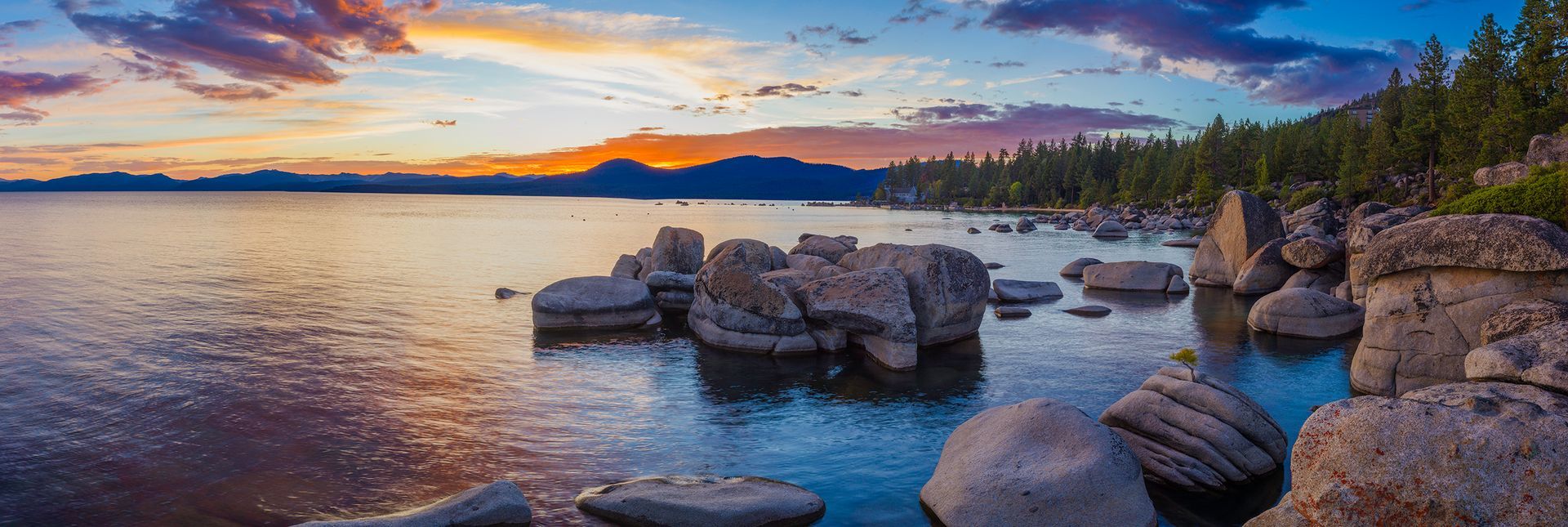 Sunset over a rocky lakeshore with calm water, colorful clouds, and snow-capped mountains in the distance