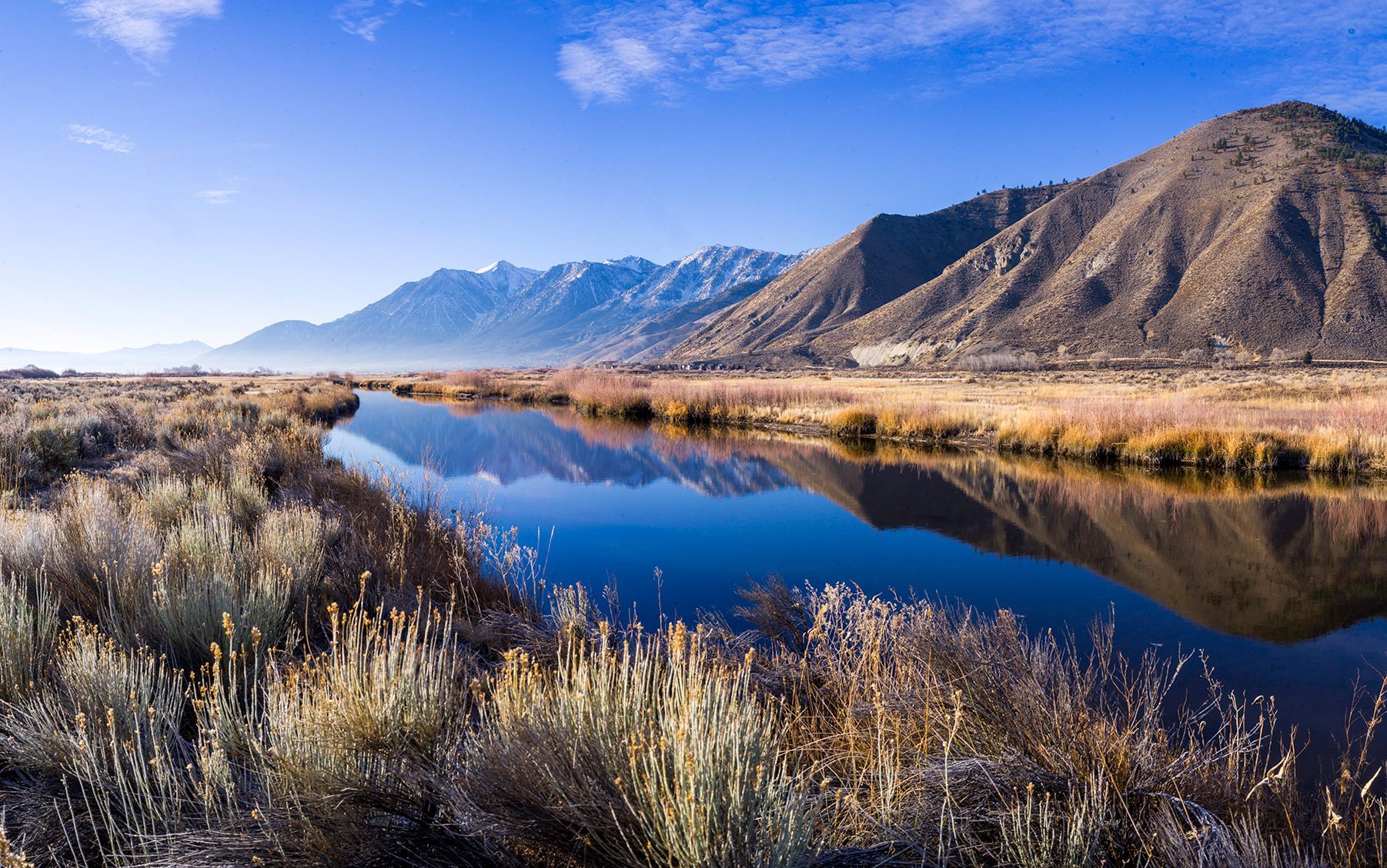 Mountain river reflecting blue sky, with golden grasses and snowless peaks in the background.